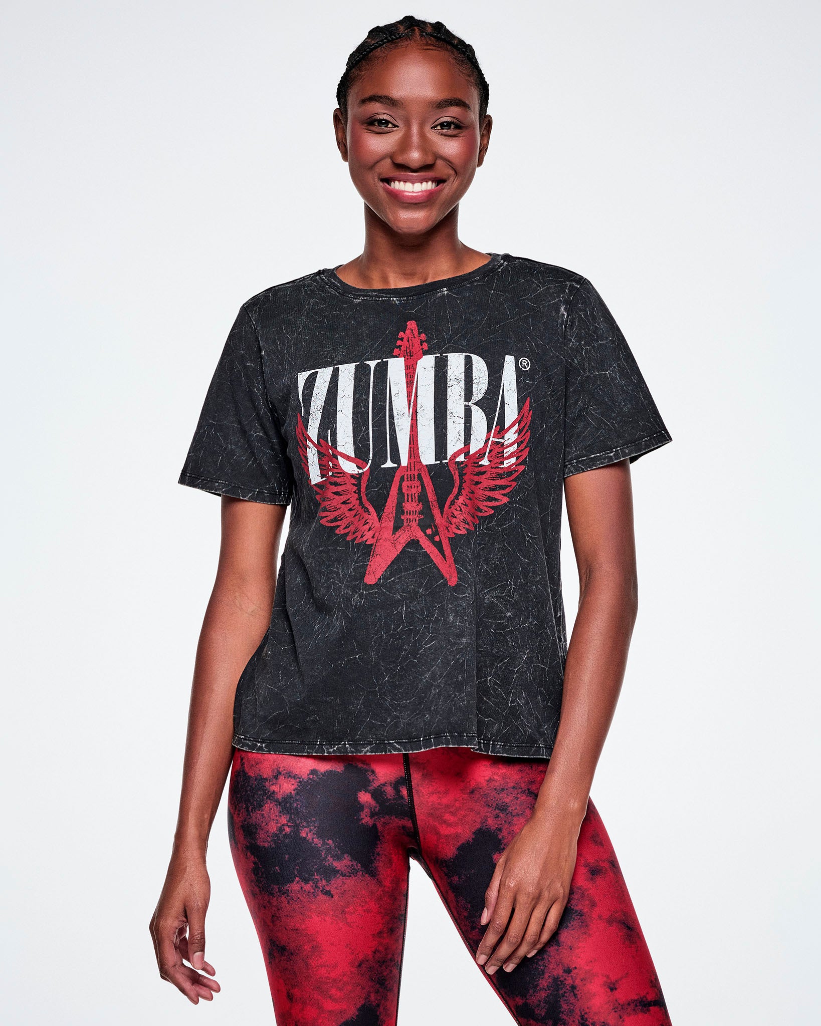 A female model wearing a black acid-washed patterned Zumba graphic tee with a red guitar with wings logo and ZUMBA in big white text, paired with red and black leggings.