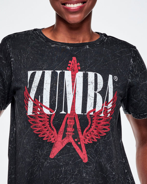 Close-up of a female model wearing a black acid-washed patterned Zumba graphic tee with a red guitar with wings logo and ZUMBA in big white text.