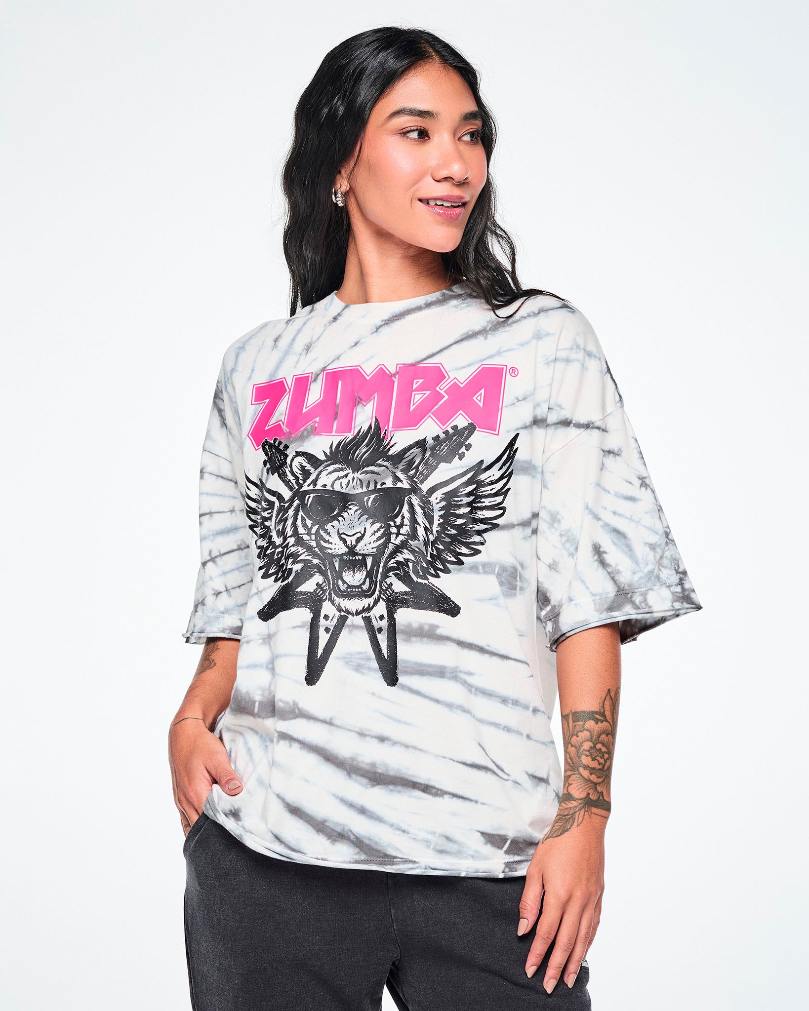 A female model wearing a white and gray tie-dye patterned graphic tee, featuring a pink 'ZUMBA' rock-like text and a black and white tiger in sunglasses with guitars and angel wings behind, paired with black pants.