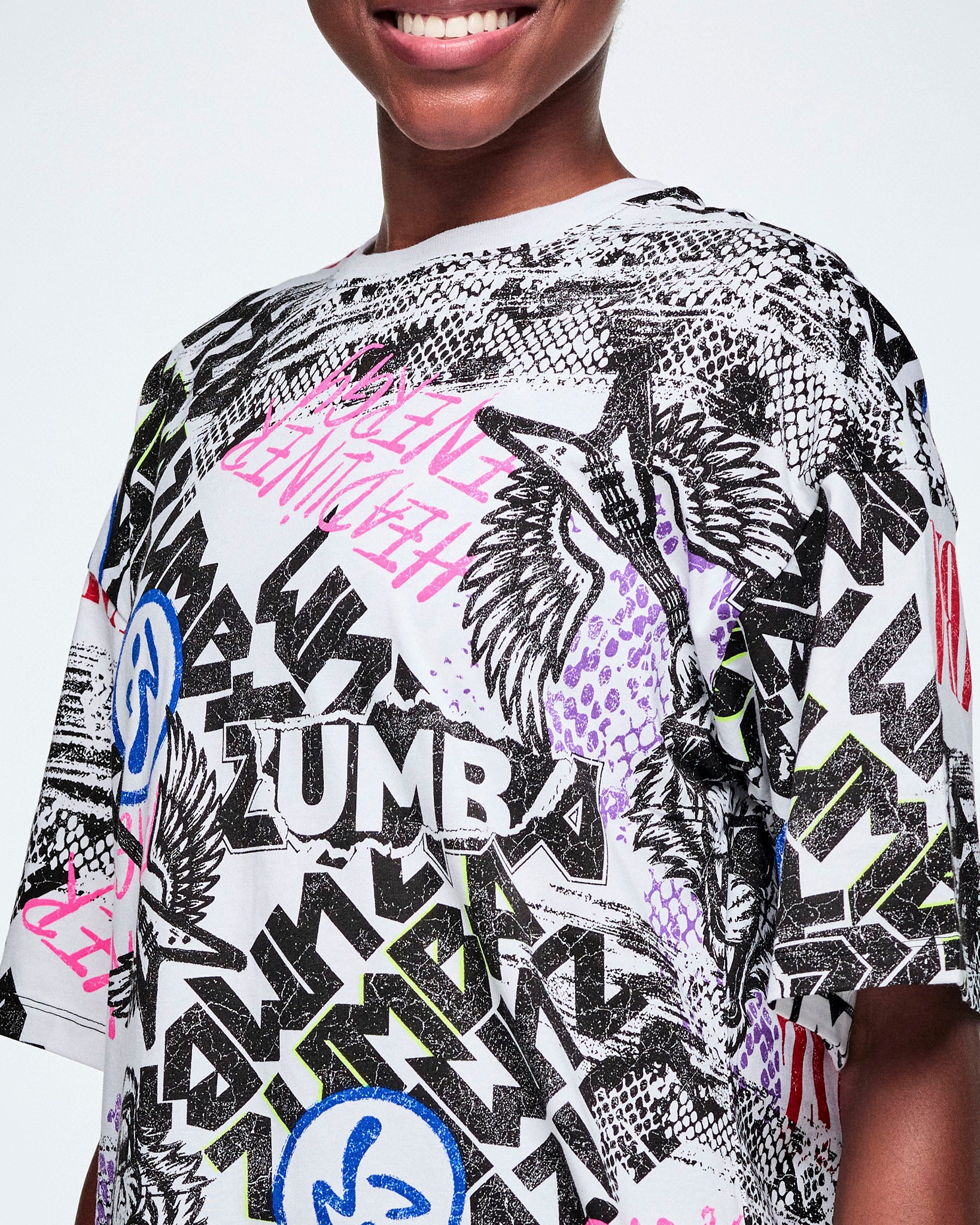 Close-up of the graphic tee with black, white, pink, and blue graffiti-style print and logos, such as blue zumbitos, a tiger wearing sunglasses, "Headliner energy, "Dance this Way," and ZUMBA in various rock and metal text, worn by a smiling female model.