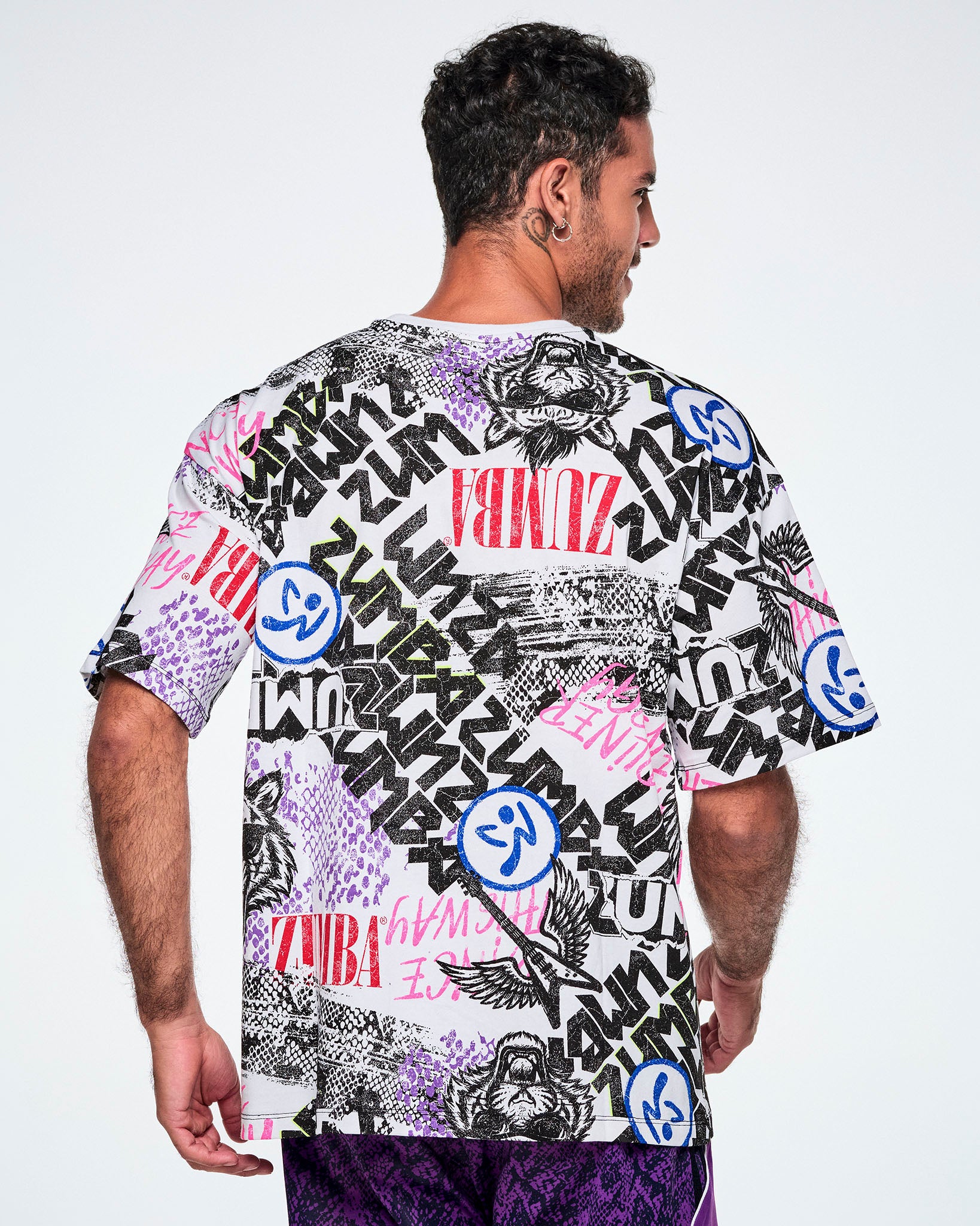 Back view of a male model wearing a graphic tee with black, white, pink, and blue graffiti-style print and logos, such as blue zumbitos, a tiger wearing sunglasses, "Headliner energy, "Dance this Way," and ZUMBA in various rock and metal text, paired with purple pants.