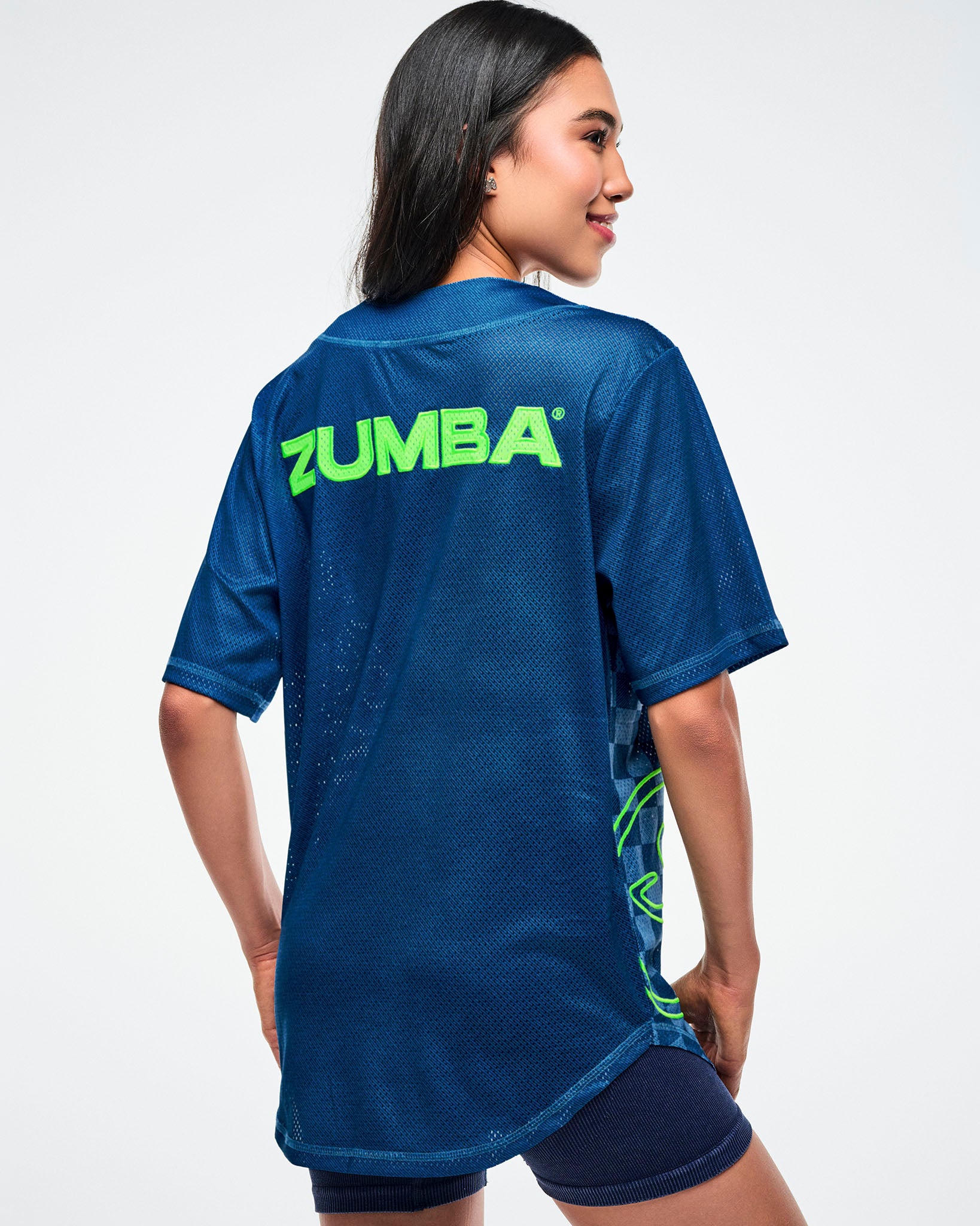 Female model, back view, wearing a blue button-up with “Zumba” in green across the upper back, paired with blue shorts.