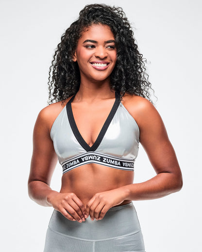 Female model wearing a silver Zumba V-neck sports bra with black trim and a black-and-white elastic band featuring repeating “ZUMBA” text paired with silver biker shorts.