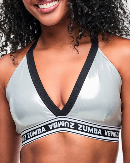 Female model wearing a silver Zumba V-neck sports bra with black trim and a black-and-white elastic band featuring repeating “ZUMBA” text.
