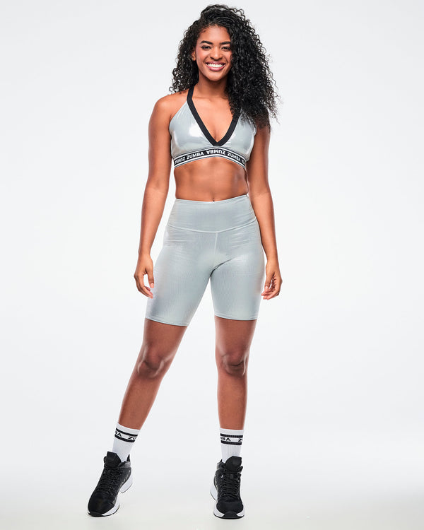 Female model wearing a silver Zumba V-neck sports bra with black trim and a black-and-white elastic band with repeating “ZUMBA” text, paired with silver biker shorts and black-and-white socks and shoes.