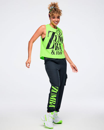 Full view of a female model wearing a neon green sleeveless Zumba tank with oversized black “ZUMBA & FLOW” text and Zumbito graphic, paired with black pants and green and white shoes.