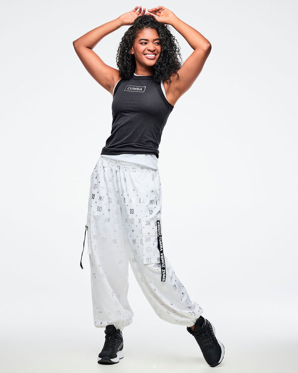 Female model wearing a black tank top with a silver 'Zumba' logo on the chest, paired with white and silver patterned cargo pants.