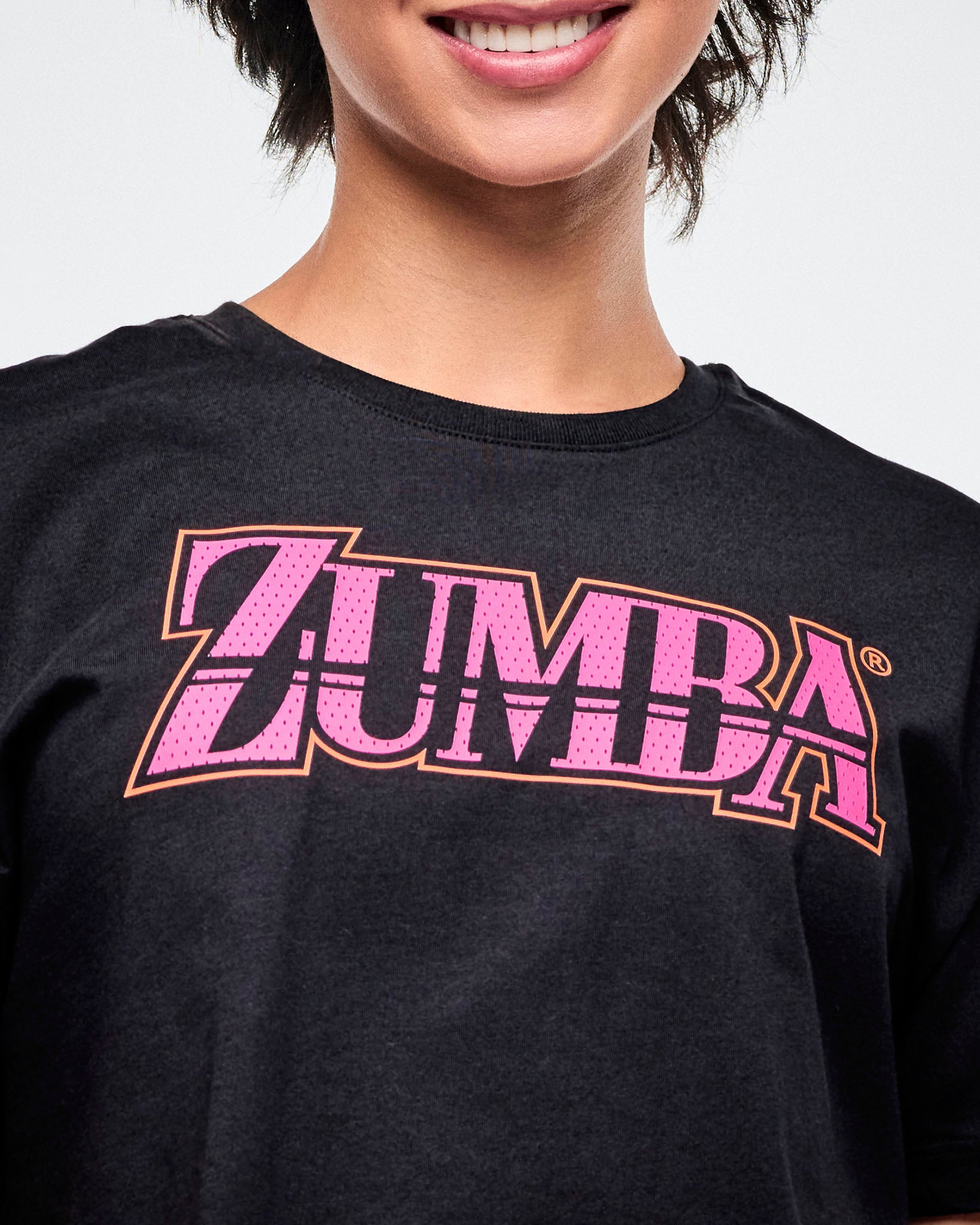 Close-up view of the female model in a black Zumba T-shirt with pink “ZUMBA” across the chest in a mesh-like pattern.