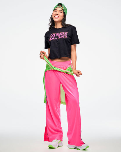 Female model wearing a backward green Zumba hat, a black Zumba T-shirt with pink “ZUMBA” across the chest, pink Zumba pants, and green-and-white shoes, with a green shirt tied around her waist.