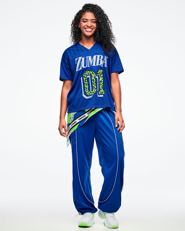 Female model wearing a blue Zumba mesh jersey with white “ZUMBA” text and a green leopard-print “01,” paired with blue-and-white pants, green-and-white shoes, and a bandana tied around her waist.