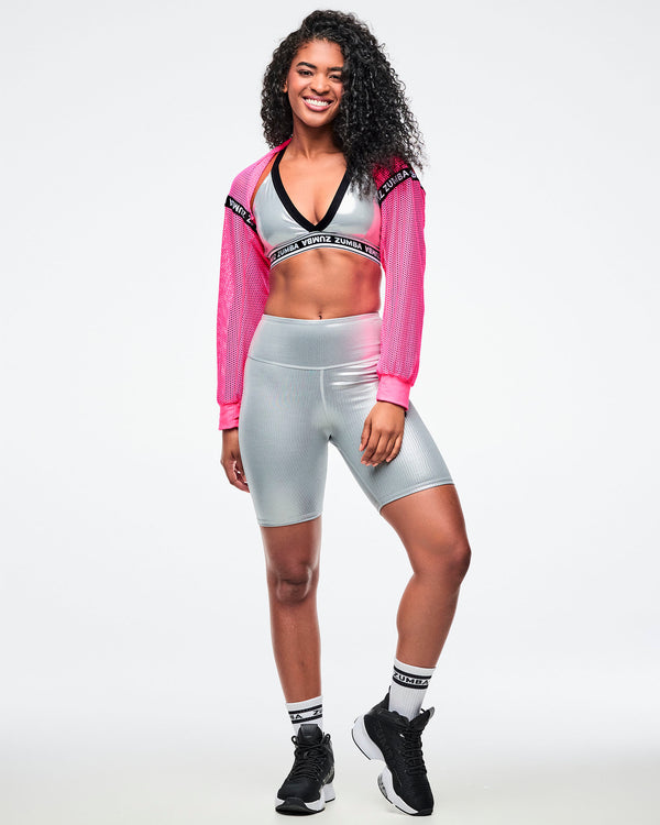 Female model wearing a pink cropped top, revealing a silver bra with matching biker shorts, paired with black-and-white socks and shoes.
