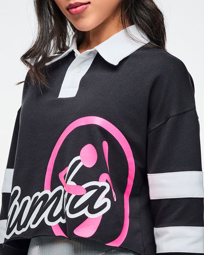 Close view of the black cropped polo with bold white “ZUMBA” text and a large pink Zumbito graphic, showcasing the white collar and white sleeve stripes.