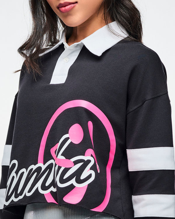 Close view of the black cropped polo with bold white “ZUMBA” text and a large pink Zumbito graphic, showcasing the white collar and white sleeve stripes.