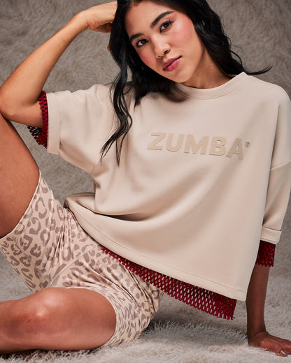 Female model seated on a shaggy cream rug in the beige drop shoulder crop top, layered over a dark red mesh top with beige leopard print biker shorts. Taupe faux fur backdrop.