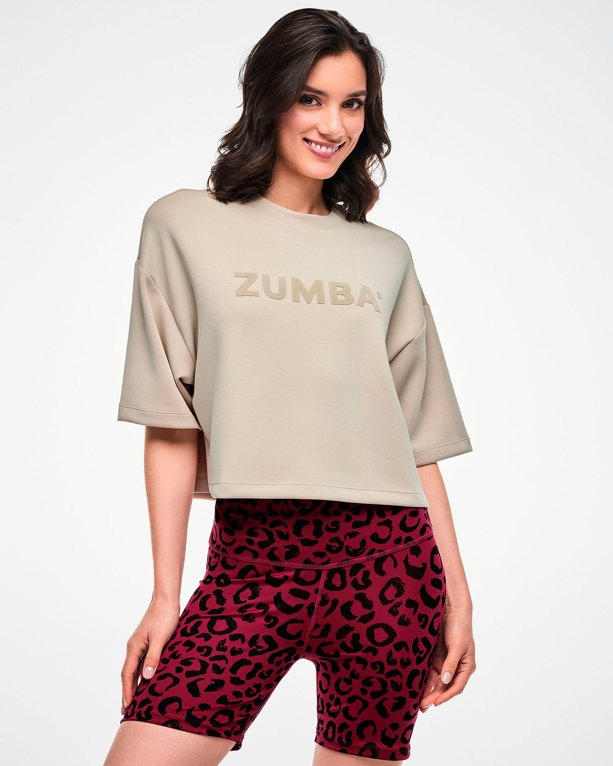 Female model in a beige oversized drop shoulder crop top with raised tonal 'ZUMBA' text across the chest, paired with dark red leopard print biker shorts.