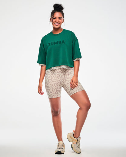 Full view of a female model in the teal green drop shoulder crop top layered over a beige mesh top, paired with beige leopard print biker shorts and beige shoes.