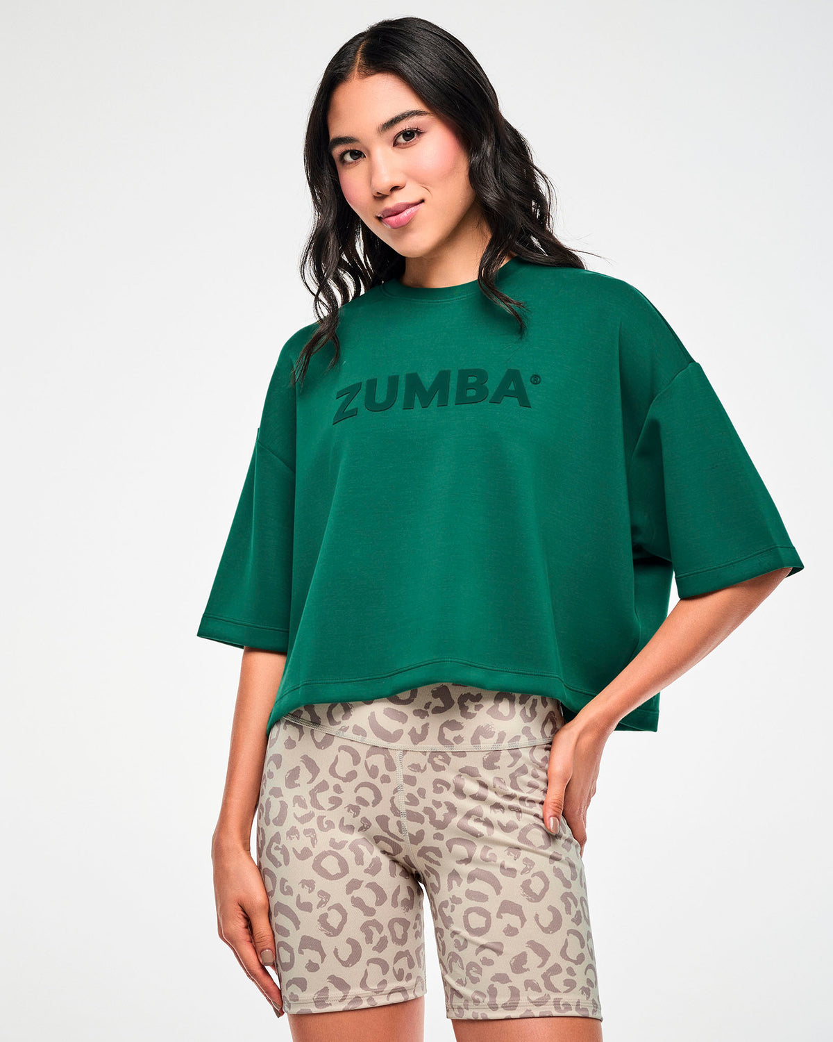 Female model in a teal green oversized drop shoulder crop top with large tonal 'ZUMBA' text across the chest, paired with beige leopard print biker shorts.