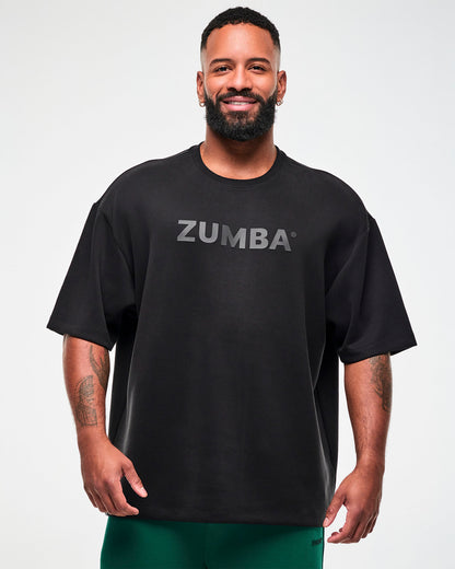 Male model in a black oversized drop shoulder crew neck top with large dark 'ZUMBA' text across the chest, paired with teal green pants.