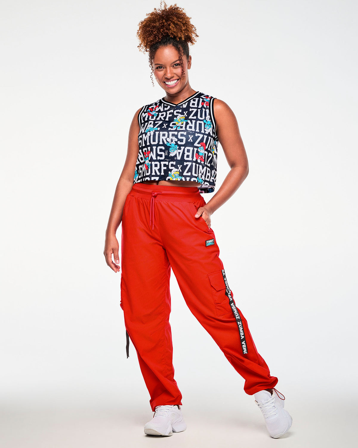 Female model with one hand in her pocket, wearing the black Smurfs character and text print crop tank with striped V-neck trim, paired with red cargo pants.