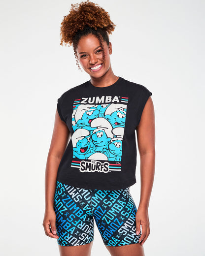 Female model in a black extended-shoulder tank with turnback cuffs, featuring white 'ZUMBA' text over a panel of smiling blue Smurfs in white hats, with 'THE SMURFS' logo below, framed by red, blue, and white stripes. Paired with blue logo print biker shorts.