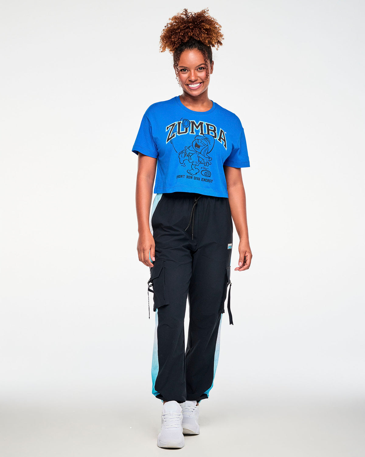 Female model in the blue Smurfette graphic crop top with arched 'ZUMBA' and 'FRONT ROW DIVA ENERGY' text, paired with black cargo pants with light blue mesh side panels and white shoes.