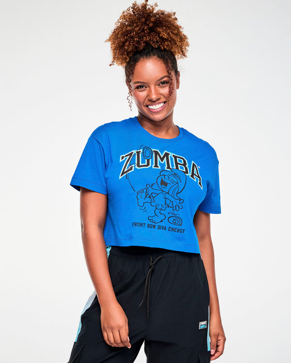 Female model in a blue relaxed crew neck crop top with the arched 'ZUMBA' and dancing Smurfette graphic and 'FRONT ROW DIVA ENERGY' text. Paired with black cargo pants with light blue mesh panels.