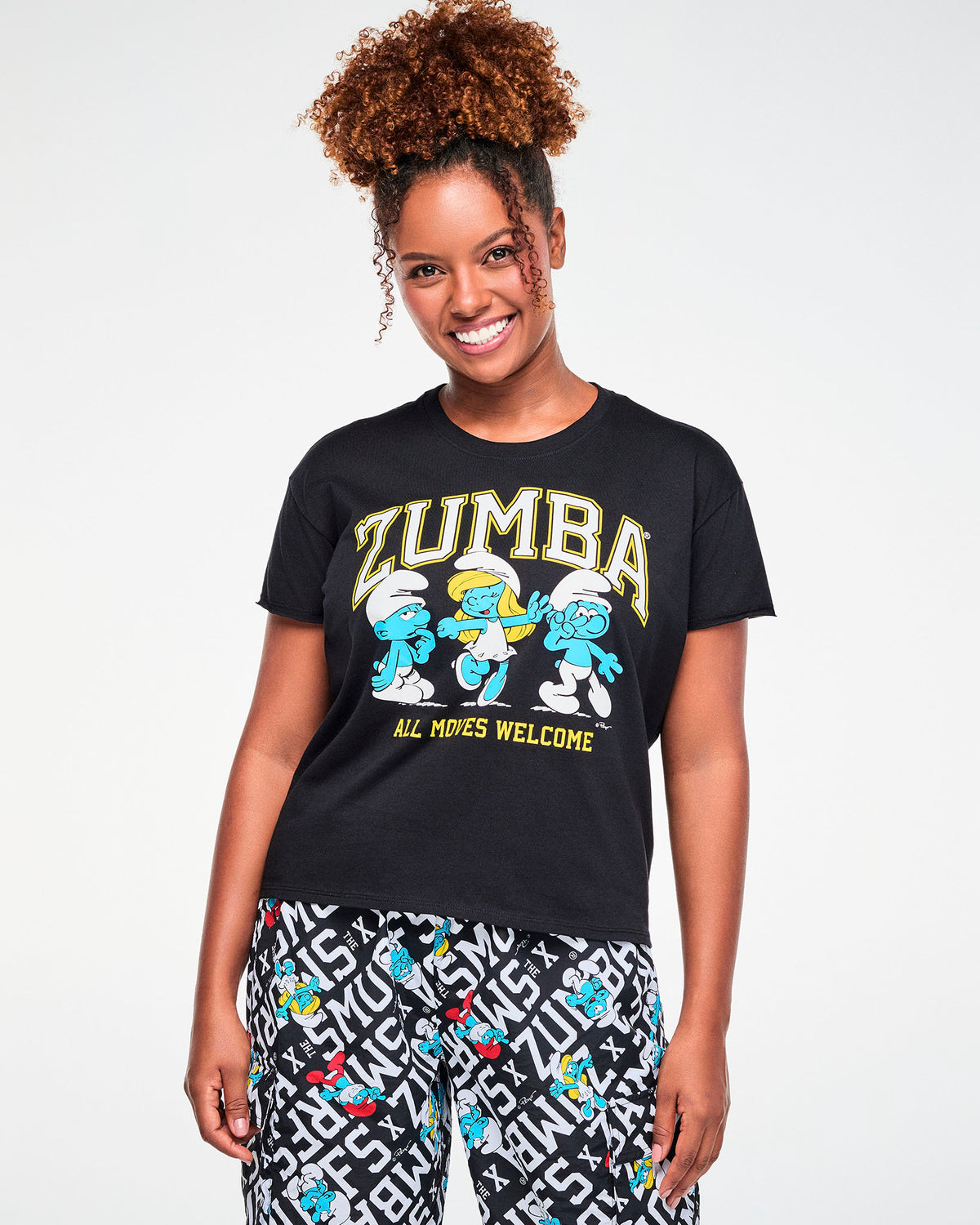 Female model in a black short-sleeve crew neck top with raw-edge sleeves, featuring arched 'ZUMBA' text, three dancing Smurfs including Smurfette, and 'ALL MOVES WELCOME' in yellow. Paired with Smurfs print cargo pants.