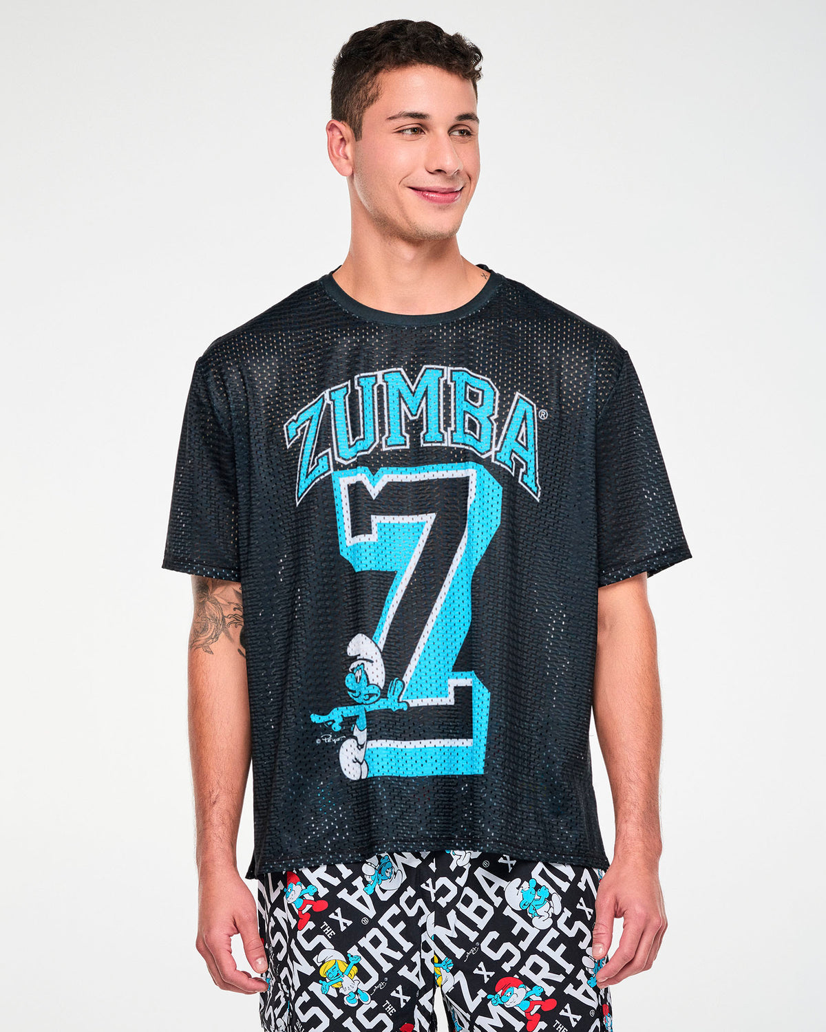 Male model in the black mesh boxy tee with the arched 'ZUMBA' text, varsity 'Z,' and leaning Smurf graphic. Paired with Smurfs print cargo pants.