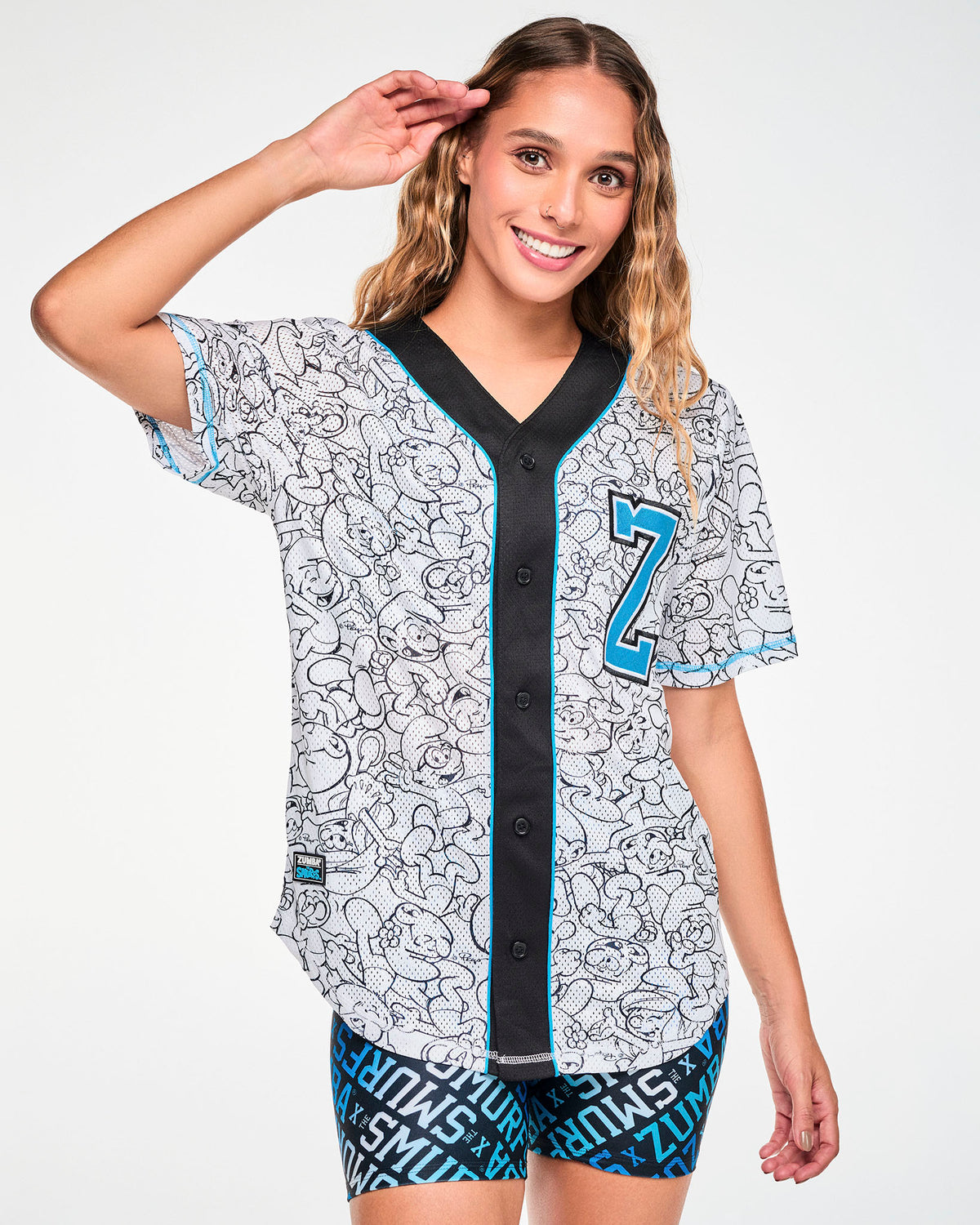 Full view of a female model in the white mesh baseball jersey buttoned up, featuring black line-drawn Smurfs all over, a black button strip (placket) with light blue piping, a light blue varsity 'Z' on the left chest, and a 'ZUMBA' X 'THE SMURFS' patch near the right hem. Paired with blue logo print biker shorts.