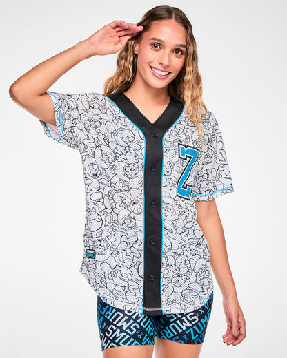 Full view of a female model in the white mesh baseball jersey buttoned up, featuring black line-drawn Smurfs all over, a black button strip (placket) with light blue piping, a light blue varsity 'Z' on the left chest, and a 'ZUMBA' X 'THE SMURFS' patch near the right hem. Paired with blue logo print biker shorts.