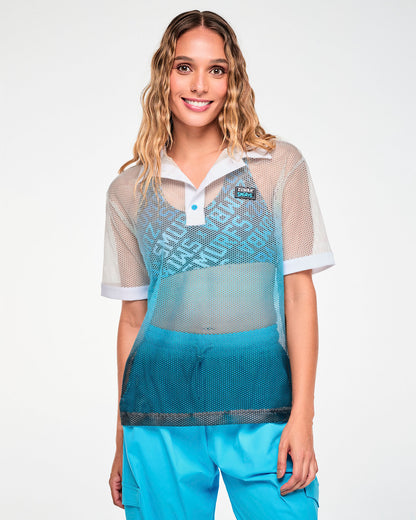 Female model in an oversized, see-through honeycomb mesh polo that fades from white at the shoulders through light blue to dark blue at the hem, with a white collar, white button strip (placket) with light blue buttons, white sleeve cuffs, and a black 'ZUMBA' X 'THE SMURFS' patch on the left chest. Worn over a blue logo print bra with aqua blue cargo pants.