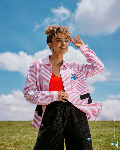 Female model in a red and white vertical striped long-sleeve button-up worn open over a red bra, with large black 'ZUMBA' letters wrapping around the lower body, paired with black tonal print pants, standing in a grassy field under a blue sky with white clouds.