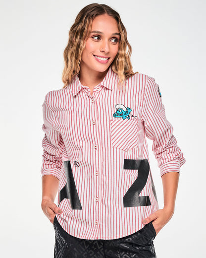 Full view of a female model in the red and white striped shirt buttoned up, showing the curved hem and the embroidered Smurf above the left chest pocket, paired with the black tonal logo print pants.