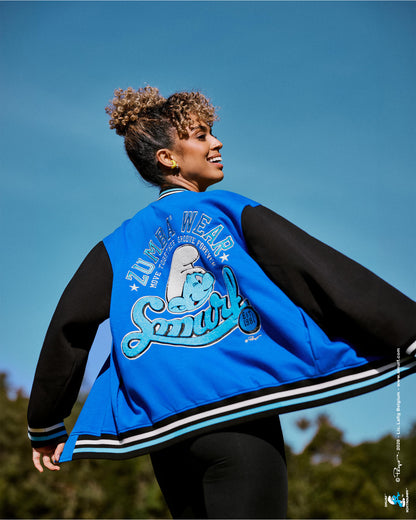 Female model twirling in a varsity jacket with a blue fleece body, black sleeves, and black, white, and light blue striped ribbed trim, showing the chenille 'ZUMBA WEAR' and 'Smurf' back graphic, with trees and blue sky behind her.