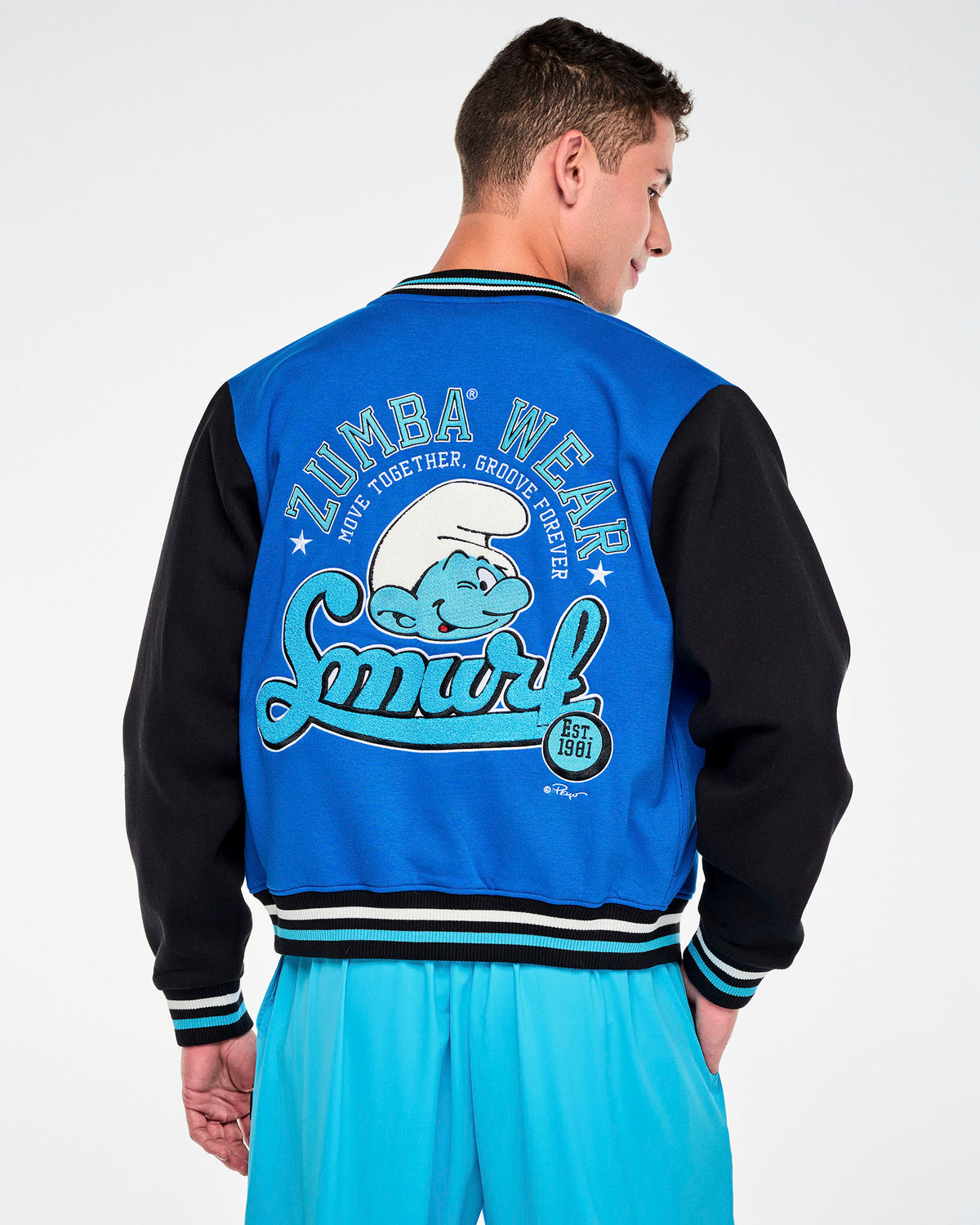 Shown from the back, a male model in the blue and black varsity jacket, with the 'ZUMBA WEAR' and 'Smurf' back graphic framed by the black sleeves and the striped ribbed hem. Paired with aqua blue cargo pants.