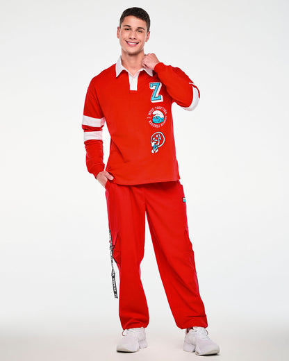 Male model holding his collar, in the red rugby polo with embroidered patches down the left front, paired with red cargo pants with black 'ZUMBA' straps.