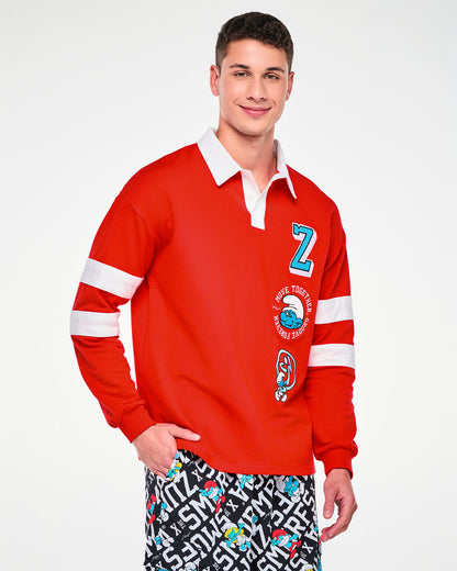 Male model in the red rugby polo, featuring the white collar, white placket, white sleeve stripes, and the embroidered 'Z,' winking Smurf, and Zumbito patches down the left front. Paired with black and white 'ZUMBA' X 'THE SMURFS' character print bottoms.