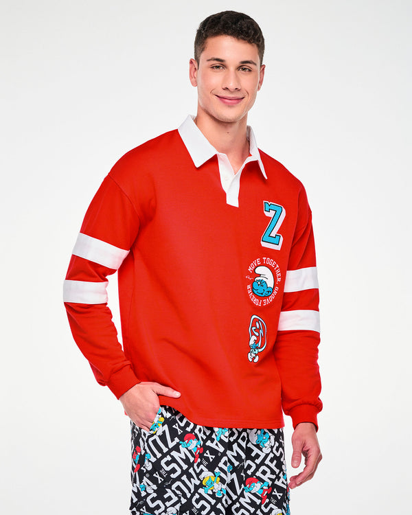 Male model in the red rugby polo, featuring the white collar, white placket, white sleeve stripes, and the embroidered 'Z,' winking Smurf, and Zumbito patches down the left front. Paired with black and white 'ZUMBA' X 'THE SMURFS' character print bottoms.