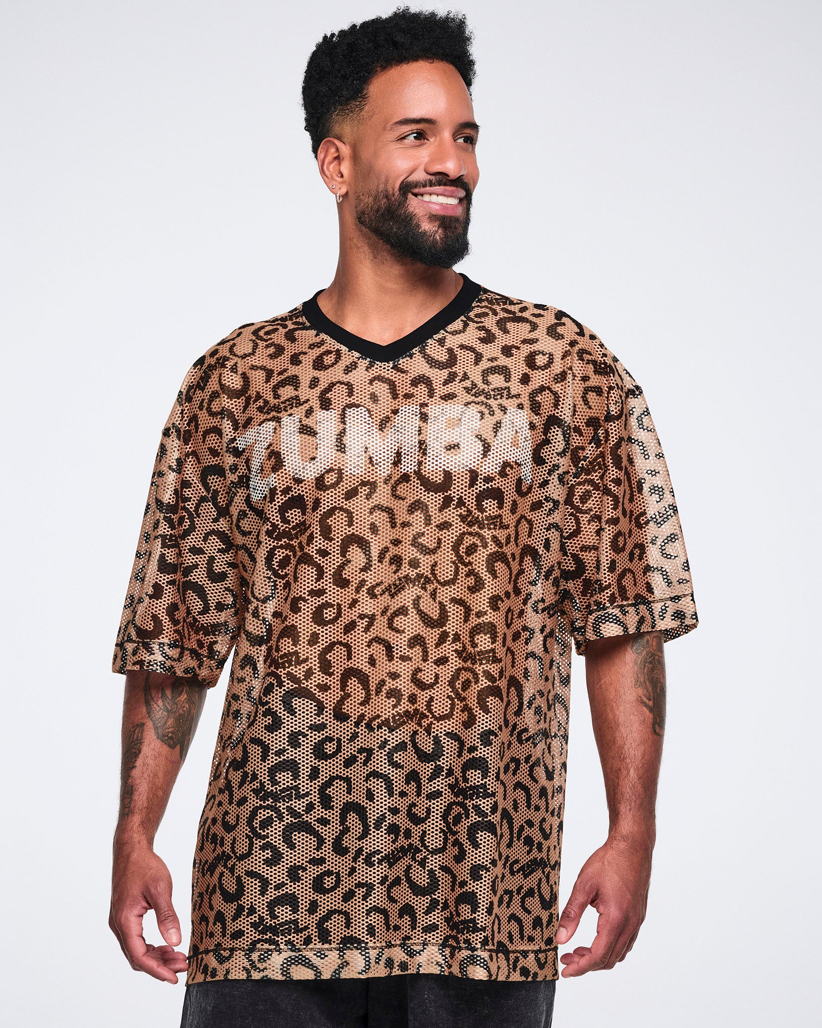 Tan leopard-print mesh top with black abstract spots, a black V-neck collar, oversized fit, and elbow-length sleeves, shown on a smiling male model wearing black long shorts.