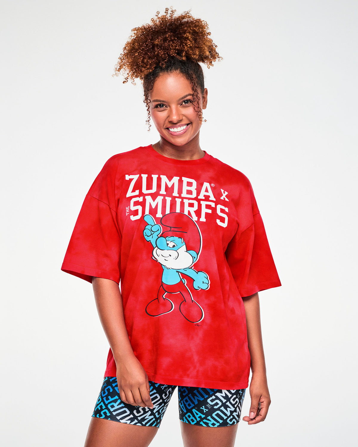 Female model in an oversized red and light cloud fabric wash tee with dropped shoulders and elbow-length sleeves, featuring white varsity 'ZUMBA' X 'THE SMURFS' text above Papa Smurf pointing one finger in the air. Paired with blue and white logo print biker shorts.