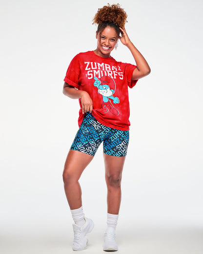 Female model with one hand in her hair, wearing the red and light cloud fabric wash tee with rolled sleeves and the white 'ZUMBA' X 'THE SMURFS' and Papa Smurf graphic, paired with blue logo print biker shorts, white socks and white shoes.