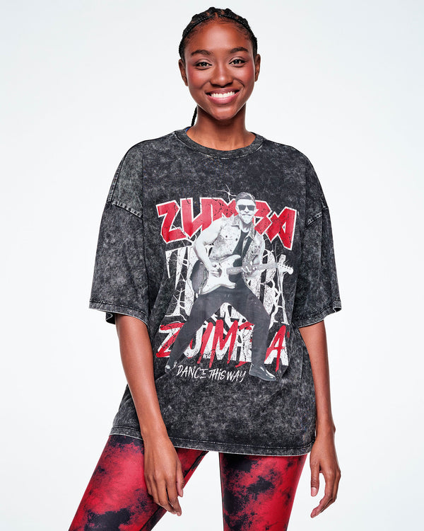 A female model wearing a black acid-wash graphic tee with Beto, the creator of Zumba, smiling in sunglasses jamming on a guitar in black and white with ZUMBA in red and white text behind him and Dance this Way in smaller white text underneath, paired with red and black leggings.