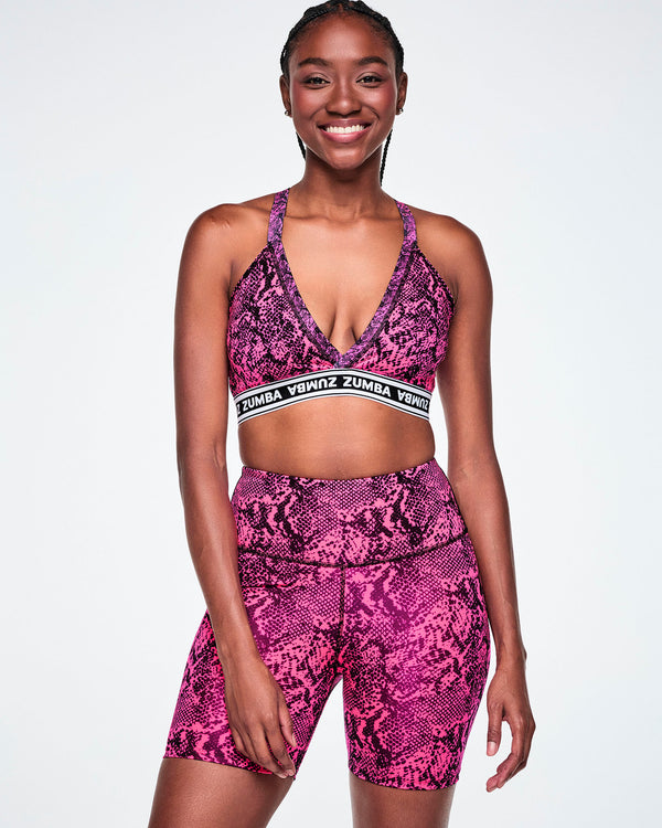 A female model wearing a pink and black snakeskin-patterned sports bra with a white and black under band printed with 'ZUMBA' in white letters and paired with matching biker shorts.