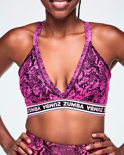 A close-up of a female model wearing a pink and black snakeskin-patterned sports bra with a white and black under band printed with 'ZUMBA' in white letters.