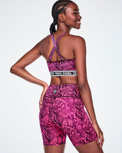 Back view of a female model wearing a pink and black snakeskin-patterned sports bra with crossover straps and a white and black under band printed with 'ZUMBA' in white letters and paired with matching biker shorts.