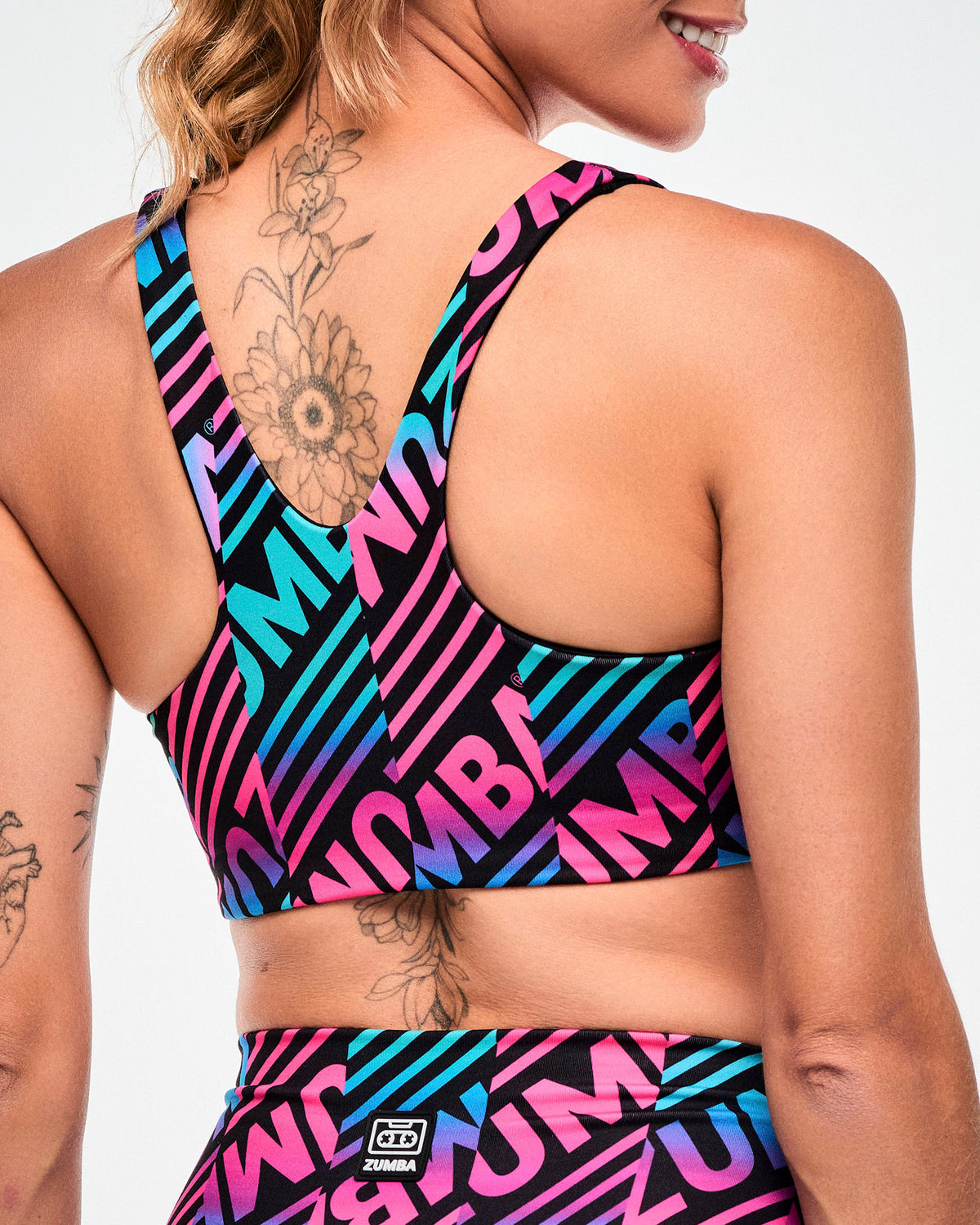 Close-up of the criss-cross back of a black sports bra with a pink, aqua, and black diagonal "ZUMBA" stripe print, paired with matching leggings with a black and white "ZUMBA" cassette tag.