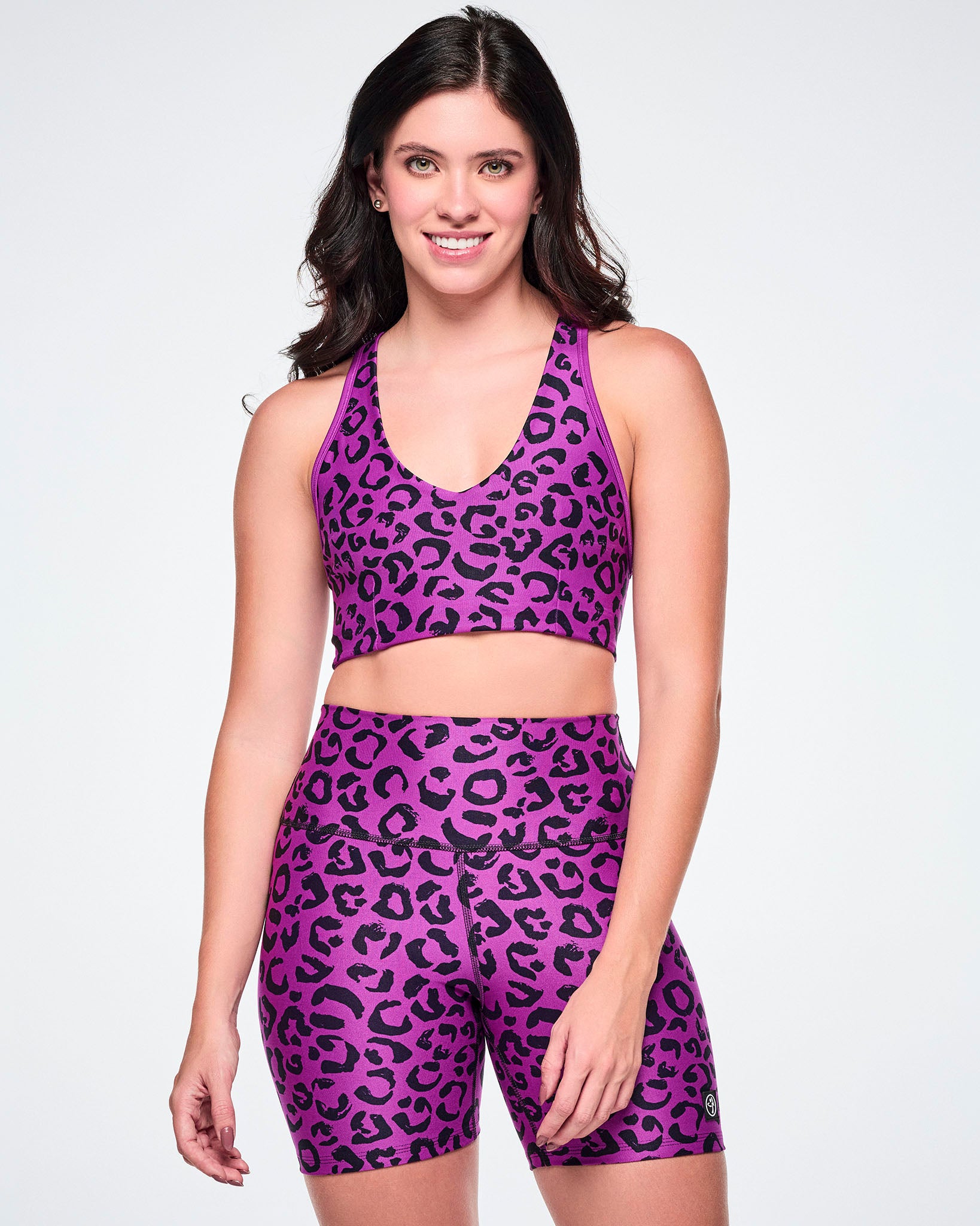 A female model wearing a matching purple and black leopard-print activewear set, featuring a reversible sports bra and high-waisted biker shorts, against a white background.