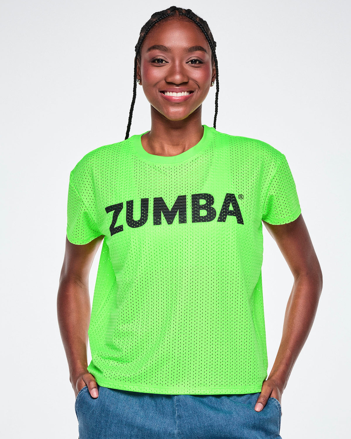 A female model wearing a bright green Zumba short-sleeve mesh top with black 'ZUMBA' text across the chest, paired with blue denim pants, against a white background.