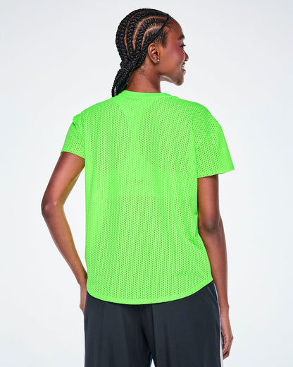 Back view of a emale model wearing a bright green Zumba short-sleeve mesh top, paired with black and blue denim pants, against a white background.