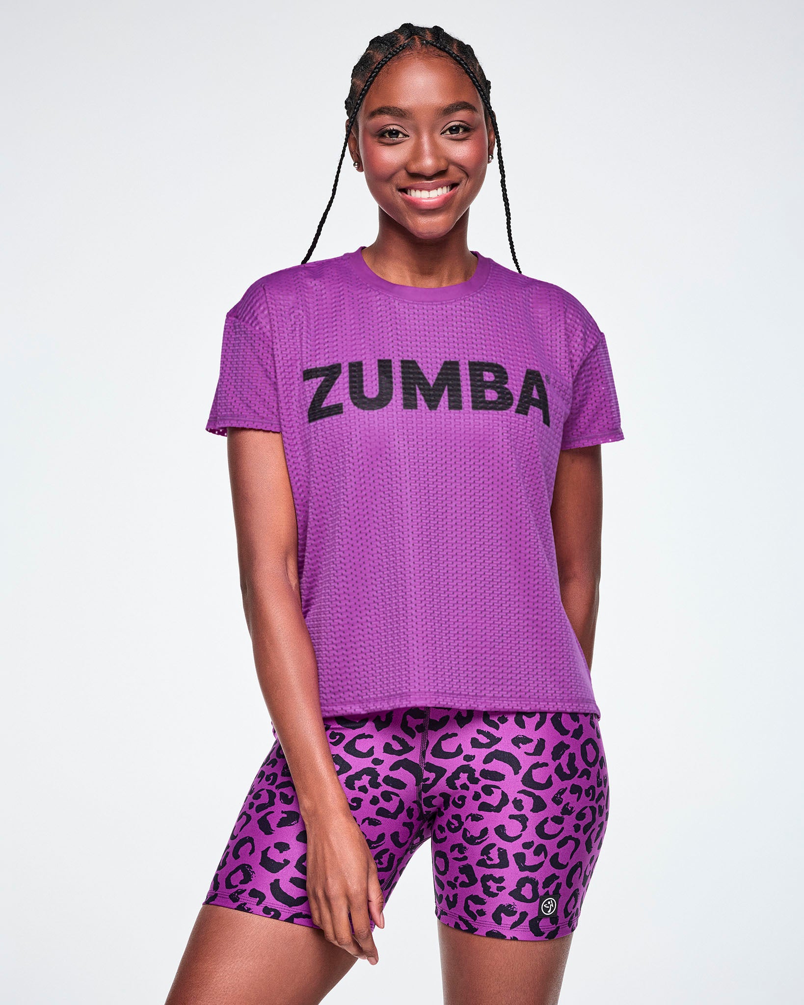 A female model wearing a purple Zumba short-sleeve mesh top with black 'ZUMBA' text across the chest, paired with purple leopard print biker shorts, against a white background.